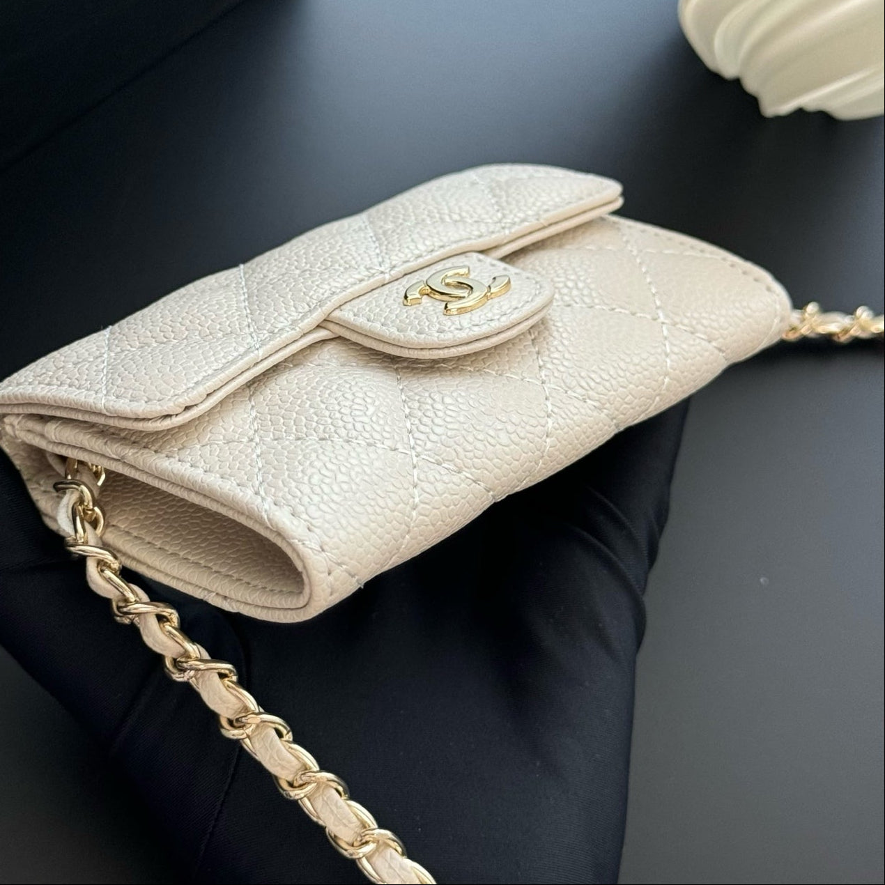 Beige handbag with gold chain held by a gloved hand on a dark background
