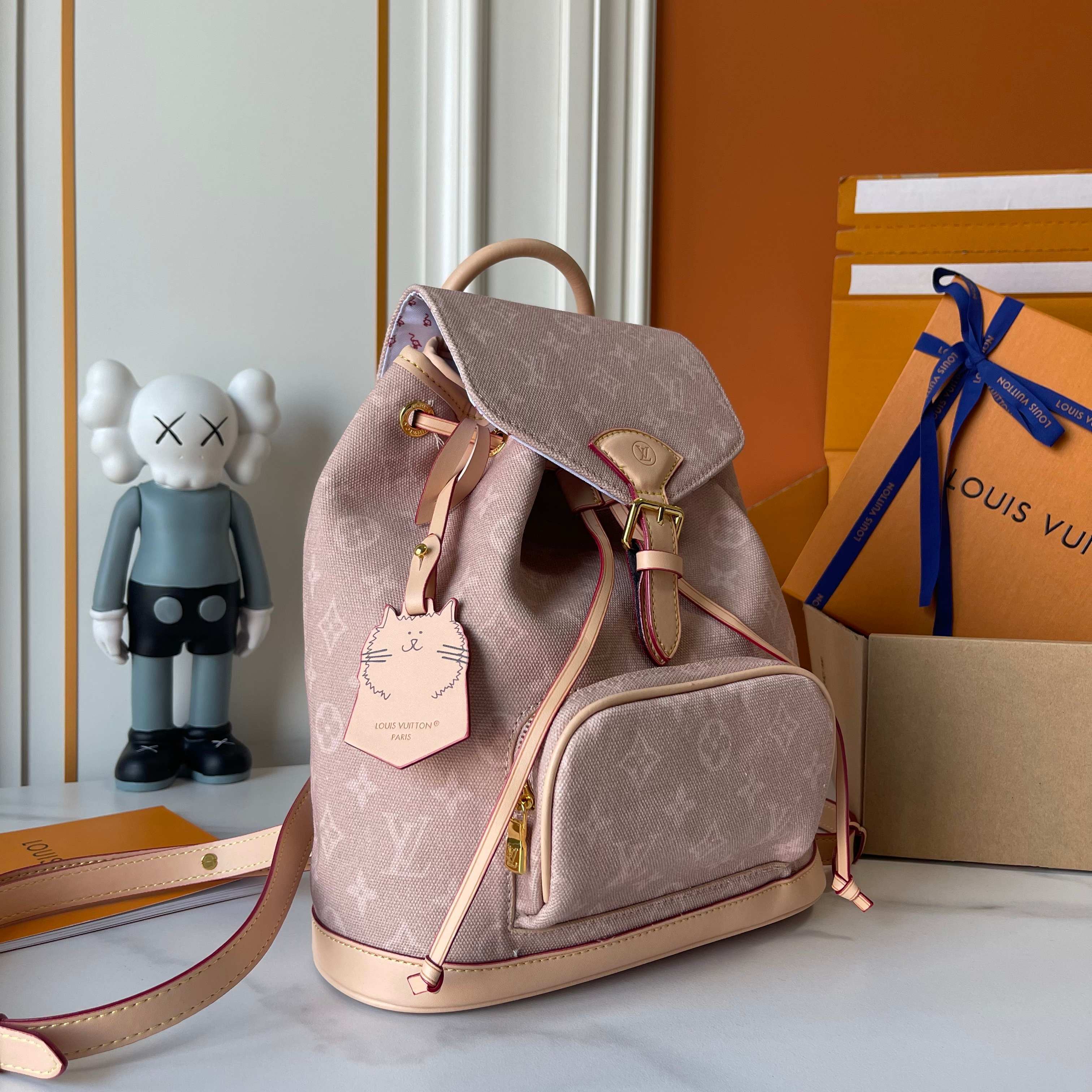 Pink backpack with gold accents on a surface with a KAWS figure and Louis Vuitton box in the background.